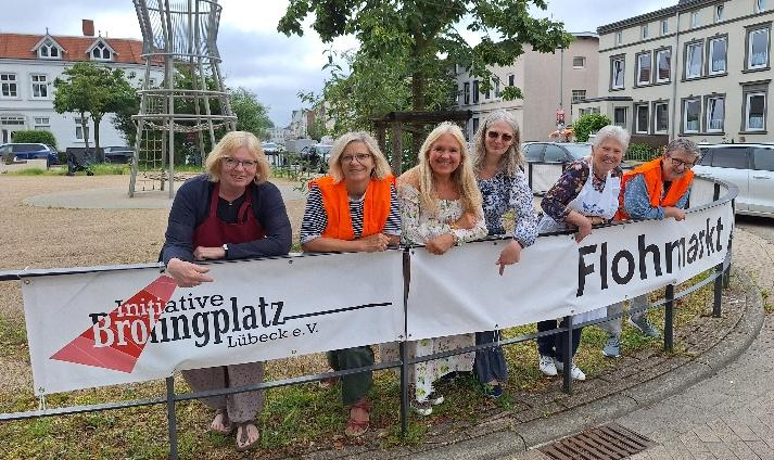 ein Team aus Ehrenamtlichen lehnt an einem Zaun, an dem Banner der Initiative Brolingplatz Lübeck eV angebracht sind, und lächeln in die Kamera